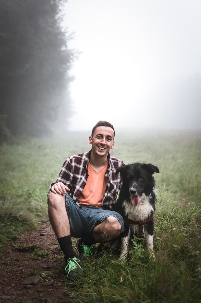 A young man crouching beside his happy border collie on a misty woodland path